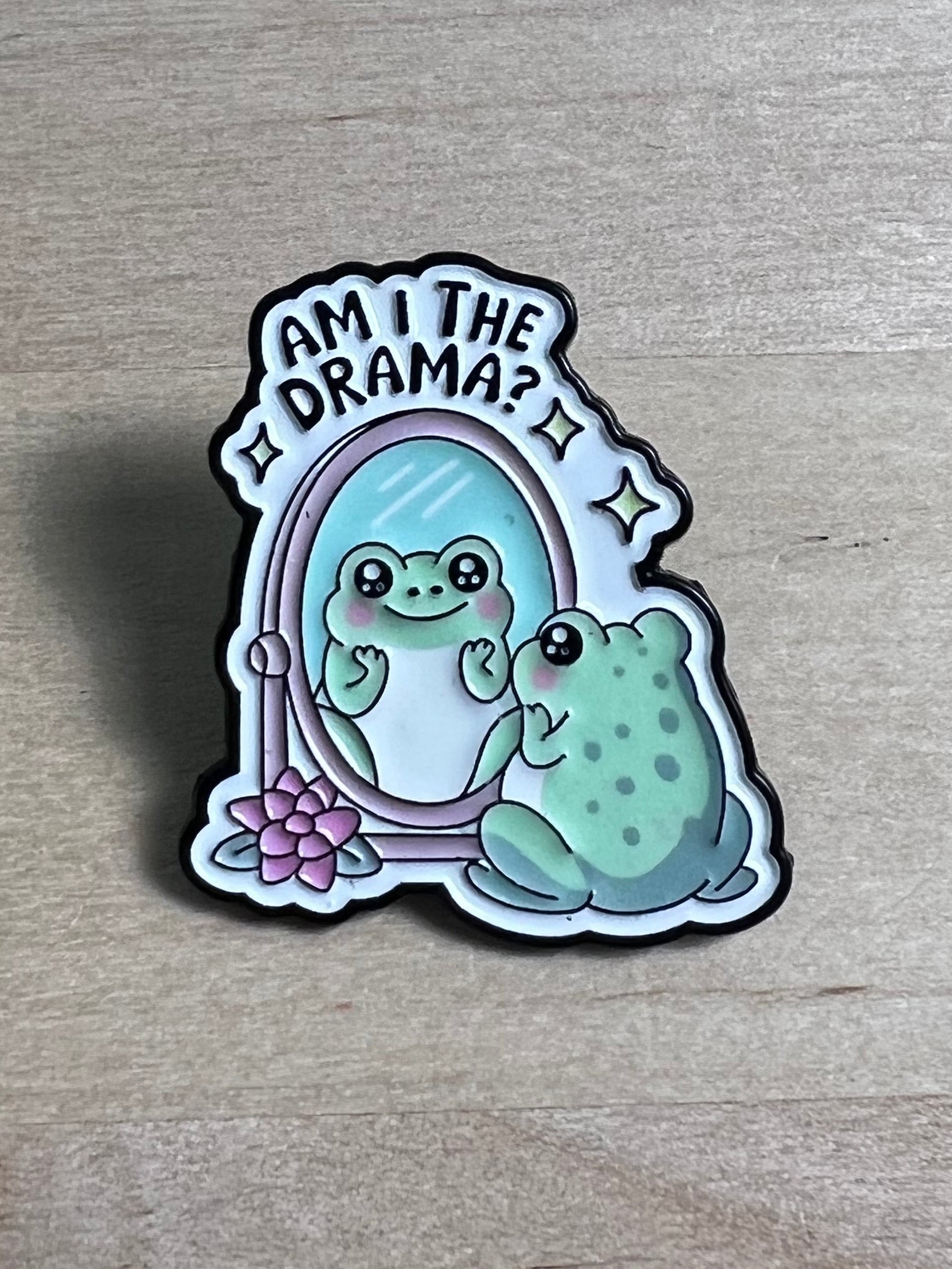 Enamel pin with a frog design and text on a wooden surface