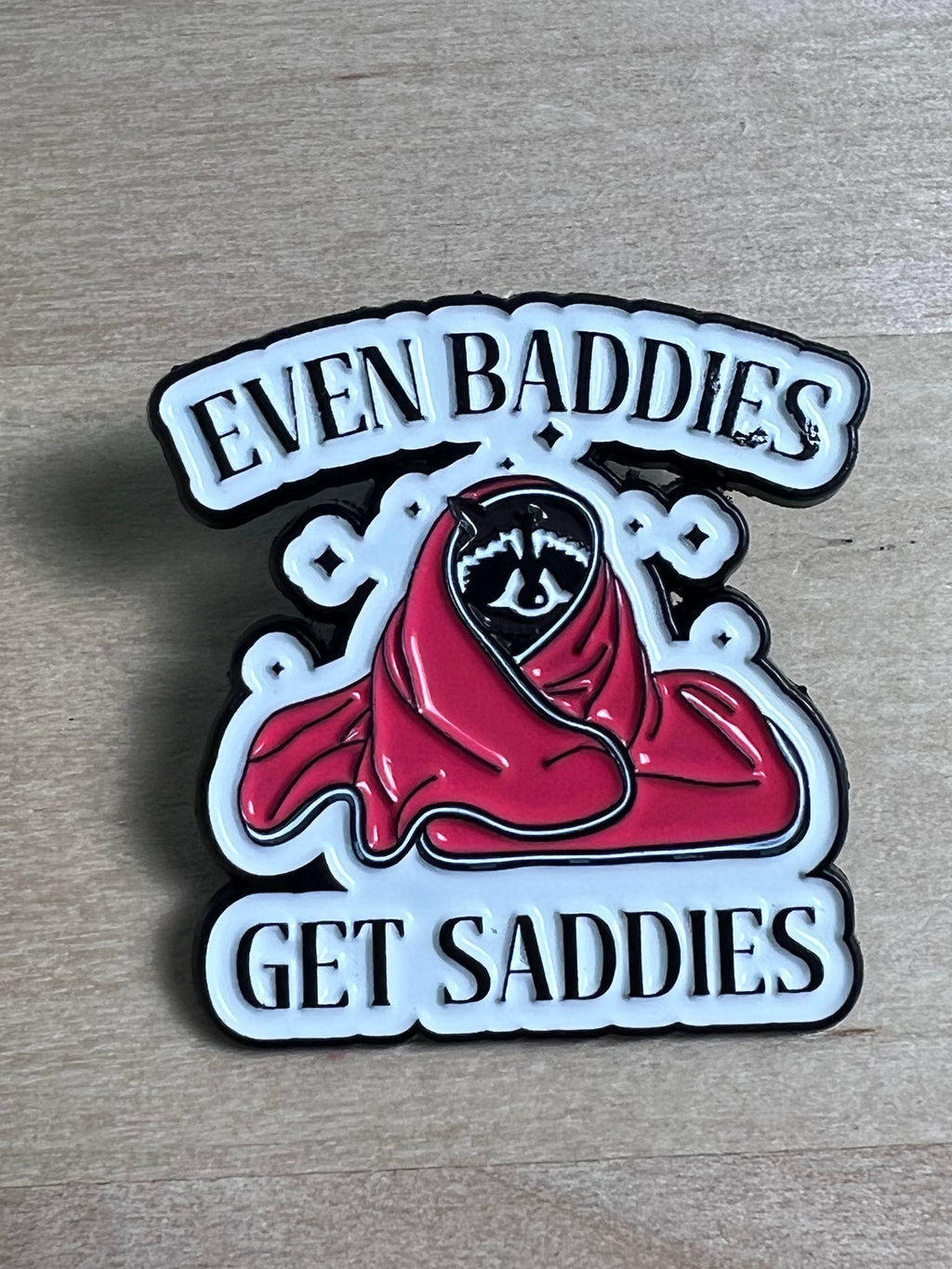 Metallic badge with red hood and text 'Even Baddies Get Saddies' on a wooden surface