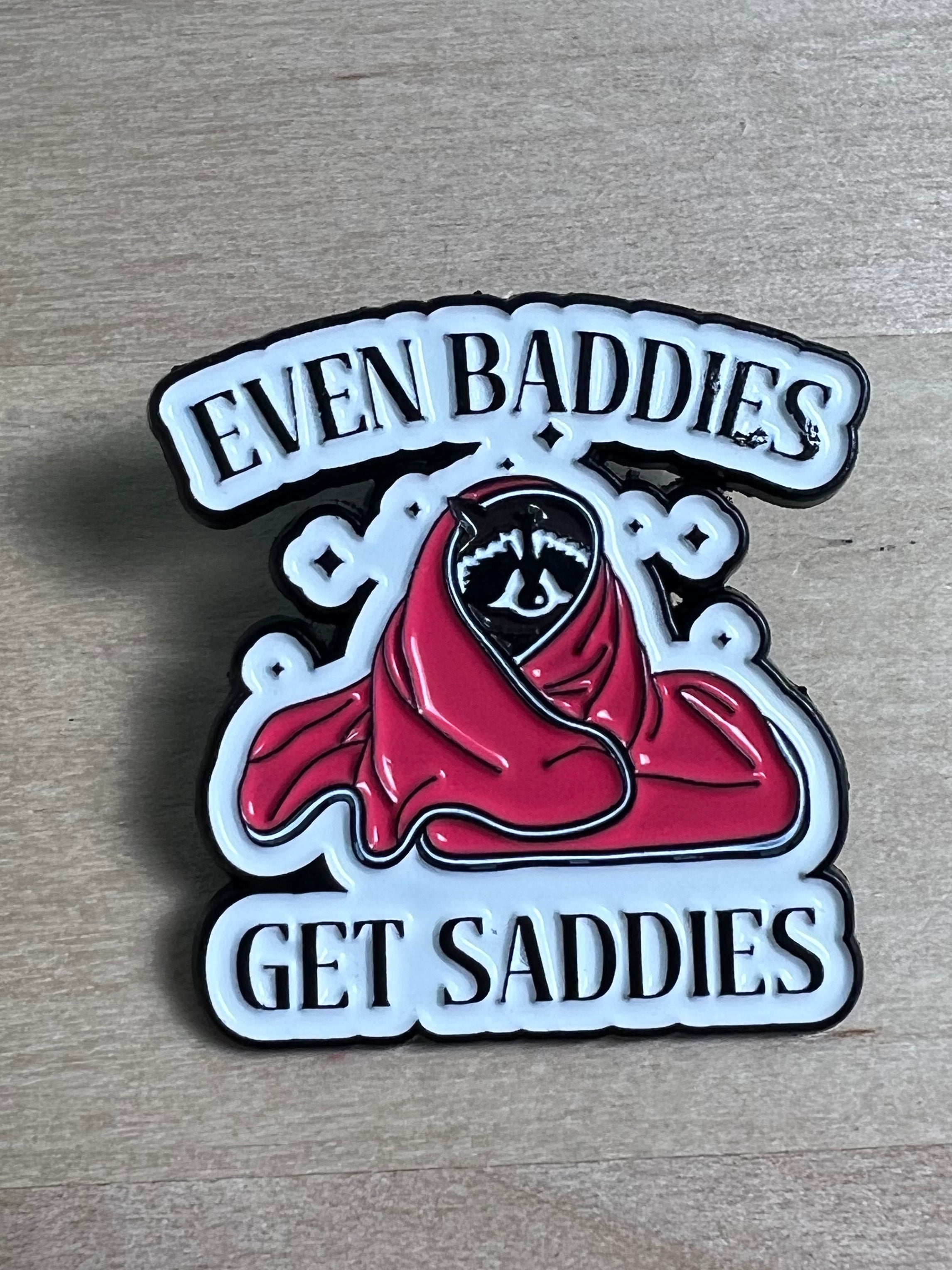 Metallic badge with red hood and text 'Even Baddies Get Saddies' on a wooden surface