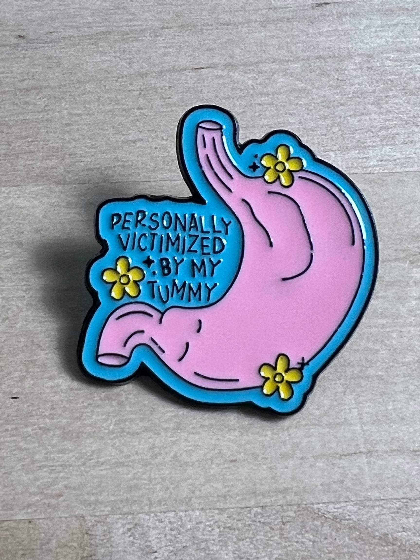 Pink and blue stomach-shaped enamel pin with text on a wooden surface